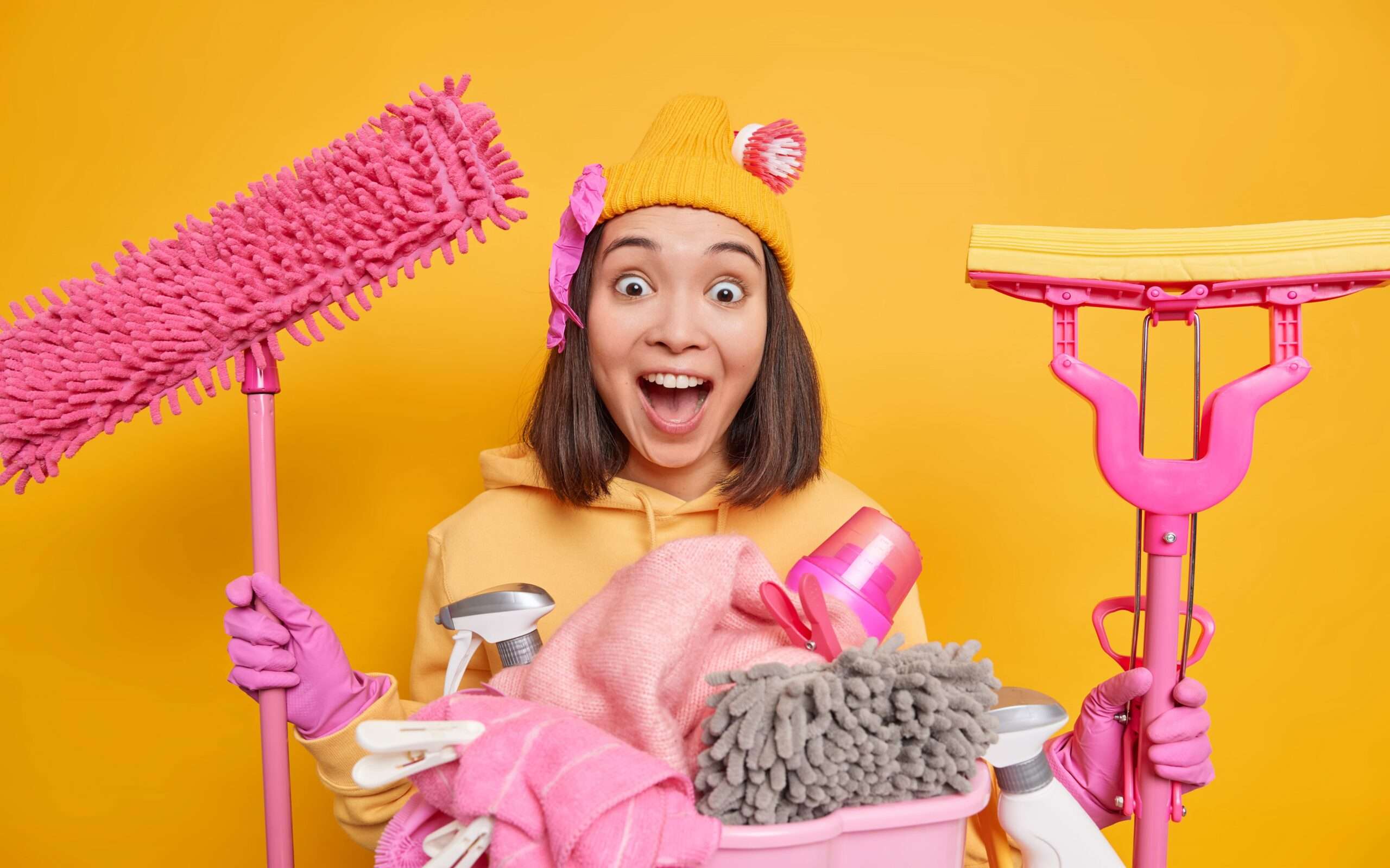 indoor shot asian woman has very surprised face keeps mouth wide opened notices dirty room going clean everything holds cleaning supplies stands near laundry basket against yellow wall min scaled